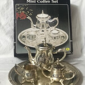 Puede incluir: Un juego de café en miniatura de plata con una tetera, una jarra de crema, un azucarero y cuatro tazas en una bandeja de plata. El juego está en su caja original, que dice "Mini Coffee Set" y tiene una imagen del juego.