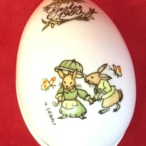 Puede incluir: Caja de porcelana blanca en forma de huevo con un diseño pintado a mano de dos conejos que se toman de la mano. Los conejos llevan ropa verde y la caja está decorada con una corona de flores y el texto "Easter 1980".