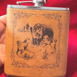 Può includere: Una fiaschetta in acciaio inossidabile rivestita in pelle color cuoio con un'incisione laser marrone scuro raffigurante un cinghiale circondato da due animali più piccoli in un bosco, racchiusa in una cornice decorativa ornata.