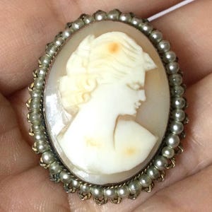 Könnte beinhalten: Ovale Kamee-Brosche mit dem Profil einer Frau, die in cremefarbenem Muschelschmuck geschnitzt ist. Die Kamee ist in einem silberfarbenen Rahmen mit einer Bordüre aus kleinen, runden, cremefarbenen Perlen gefasst. Ein klassisches Schmuckstück.