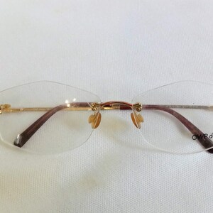 Puede incluir: Gafas sin montura con montura de metal dorado y patillas marrones. Las lentes son transparentes y tienen una forma geom&eacute;trica sutil. El texto "OWP design" est&aacute; impreso en la lente inferior derecha.