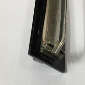 May include: Close-up of a black and silver clipboard clip. The clip has a black frame and a silver metal lever and clasp. The background is white.