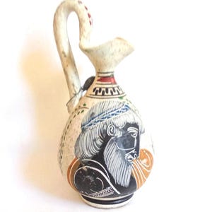 May include: A ceramic pitcher with a curved handle and a spout. The pitcher is painted with a design of a man's face in black and white, with blue and orange accents. The handle has red and white details.