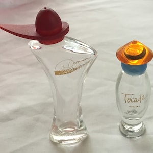 Pode incluir: Dois frascos de perfume em miniatura. O frasco à esquerda é de vidro transparente com uma tampa vermelha e a palavra "Donna" em letras douradas. O frasco à direita é de vidro transparente com uma tampa azul e uma tampa laranja em forma de chapéu. O frasco está etiquetado com "Tocade" em letras douradas.