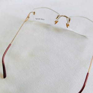 Puede incluir: Gafas sin montura con montura de metal dorado y lentes transparentes. Las patillas tienen un acabado burdeos en los extremos. Las gafas est&aacute;n dise&ntilde;adas con una est&eacute;tica minimalista.