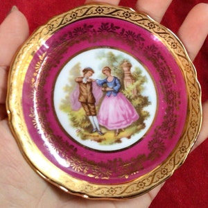 Puede incluir: Un pequeño plato de porcelana rosa y dorado con una escena romántica de una pareja en un jardín. El plato tiene un borde dorado y diseños florales intrincados.