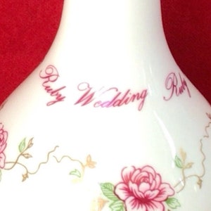 May include: White ceramic vase with a floral design and the words "Ruby Wedding" in pink script. The vase features pink roses, green leaves, and brown ribbon accents, suitable for home decor.