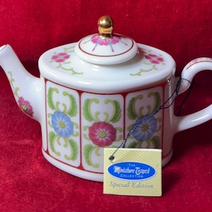 Puede incluir: Una tetera de cerámica blanca con un diseño floral en rosa, azul y verde, acentuada con ribetes rojos. La tapa está rematada con un pomo dorado. La etiqueta dice "The Miniature Teapot Collection Special Edition."