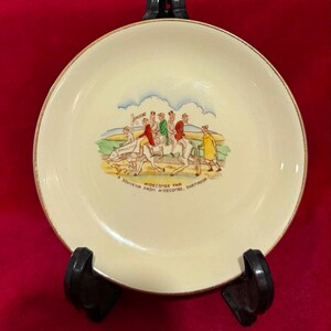Könnte beinhalten: Ein cremefarbener Keramiktarbener Keramikteller mit goldfarbenem Rand und einer bunten Illustration von Menschen auf Pferden und einem Hund. Unter der Szene steht in schwarzer Schrift WIDECOMBE FAIR und A SOUVENIR FROM WIDECOMBE, DARTMOOR auf dem Teller.