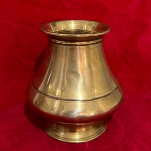 May include: A polished brass vessel featuring a wide, rounded body that tapers toward a flared rim and rests on a circular pedestal base. The metallic surface displays a warm, golden hue with subtle horizontal lines encircling the neck and base.