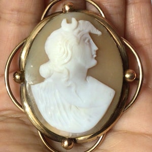 Pode incluir: Broche de camafeu oval com um perfil branco de mulher com toucado, sobre um fundo bege. O camafeu é emoldurado por uma borda de metal dourado e detalhes decorativos.