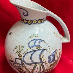 May include: A cream-colored ceramic pitcher with a round body and a curved handle. The pitcher is decorated with a nautical scene featuring a ship with blue and brown sails. The rim is adorned with blue and yellow spiral designs.
