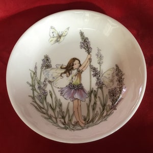 Puede incluir: Un plato de cerámica blanca con un diseño de hada de lavanda. El hada sostiene un tallo de lavanda y hay mariposas volando a su alrededor. El plato es perfecto para guardar objetos pequeños como joyas o baratijas.