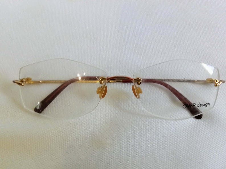 Puede incluir: Gafas sin montura con puente y varillas doradas y patillas marrones. Las lentes son transparentes y tienen un dise&ntilde;o geom&eacute;trico sutil. Las almohadillas nasales son de color dorado claro. El texto "OWP design" es visible.