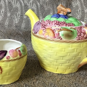 Peut inclure: Une théière et un sucrier en céramique jaune avec un motif de panier de fruits. La théière a une poignée en forme de branche d'arbre et un couvercle avec un motif de fruits. Le sucrier a un motif de fruits autour du bord.