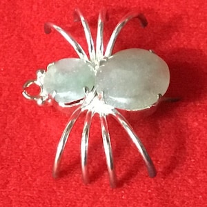 Può includere: Un ciondolo a forma di ragno in argento con due pietre di giada verde chiaro. Il ragno ha otto zampe e un corpo dettagliato.