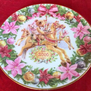 May include: A white porcelain Christmas plate with a gold rim, featuring a carousel with three teddy bears riding a horse. The plate is decorated with a green wreath of holly, poinsettias, and ornaments. The year 2000 is printed in the center of the plate.