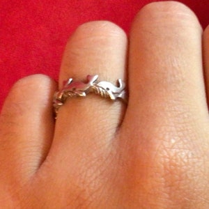 May include: A silver ring featuring a design of interwoven branches. The ring is worn on a person's finger.