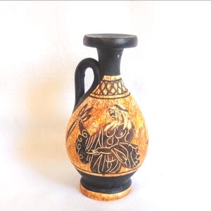 May include: A small, decorative ceramic vase with a black handle, rim, and base. The vase features a tan body with a black illustration of a figure holding a trident. The vase is approximately 6 inches tall.
