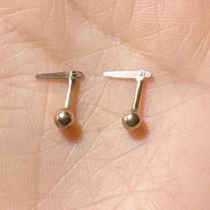 May include: Two small, gold-colored cufflinks with a ball at the bottom and a flat, rectangular top. The cufflinks are displayed on a light skin-toned background.