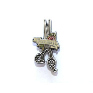 May include: Silver-toned enamel pin featuring a pair of scissors with a red rose and green leaves. A banner with the text "STAY SHARP" is wrapped around the scissors.