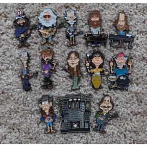 Pode incluir: Uma coleção de alfinetes de esmalte coloridos com caricaturas de músicos tocando vários instrumentos. Os alfinetes retratam membros da banda com guitarras, teclados, baterias e microfones.