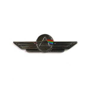 Könnte beinhalten: Eine schwarze Metallnadel mit silberner Oberfläche. Die Nadel zeigt ein kreisförmiges Design mit einem schwarzen Dreieck und einem Regenbogenprisma im Inneren. Der Kreis ist von zwei Flügeln umgeben.