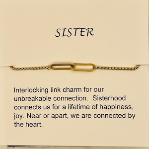 May include: A gold interlocking link charm necklace with a white card that reads "SISTER. Interlocking link charm for our unbreakable connection. Sisterhood connects us for a lifetime of happiness, joy. Near or apart, we are connected by the heart."