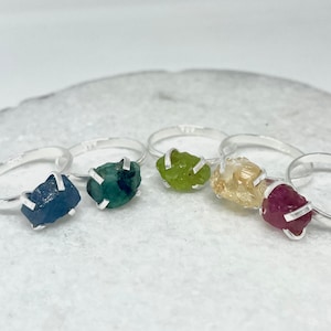May include: Five silver rings with different colored gemstones. The rings feature a blue, green, green, yellow, and red gemstone.