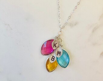 May include: A silver necklace with a delicate chain, featuring four colorful, faceted gemstone pendants in pink, yellow, and teal. Each pendant is leaf-shaped and has a silver initial charm. The initials are K, M, and B.