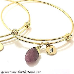 May include: Two gold bangle bracelets with charms. The charms are raw gemstones, each representing a different birth month. The bracelets have initial charms with the letters "W" and "N".