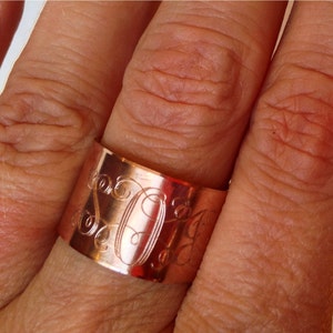 May include: A wide copper band ring with an engraved monogram. The ring is worn on a finger.