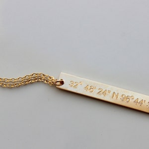 May include: Gold necklace with a long, thin bar pendant engraved with the coordinates "32&deg; 48' 24" N 96&deg; 44' 36" W".