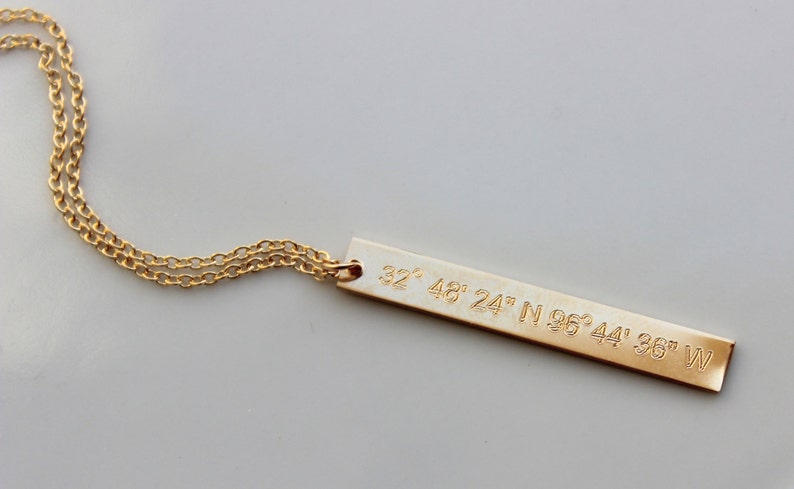 May include: Gold necklace with a long, thin bar pendant engraved with the coordinates "32&deg; 48' 24" N 96&deg; 44' 36" W".