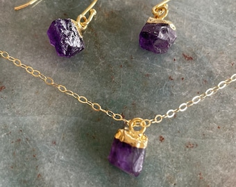 May include: A gold-colored necklace and matching earrings set. Each piece features a raw, deep purple amethyst crystal. The crystals are set in gold-colored metal. The earrings have a hook closure.