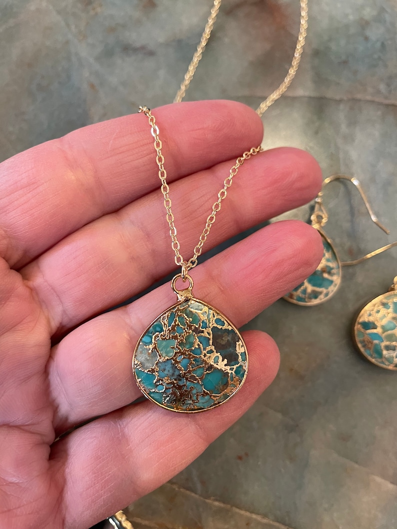 May include: A gold chain necklace with a teardrop-shaped pendant made of turquoise and gold. The pendant has a marbled pattern of blue, green, and brown. The necklace is paired with matching earrings.