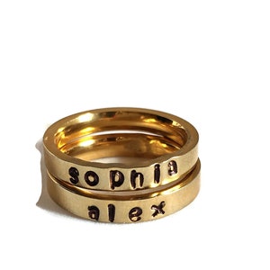 May include: Two gold-colored rings stacked on top of each other. The top ring has the word "sophia" stamped on it in black letters. The bottom ring has the word "alex" stamped on it in black letters. The rings are on a white background.