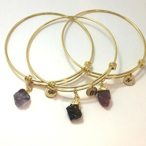 May include: Three gold bangle bracelets with charms. The charms are small, rough-cut gemstones in purple, red, and black. Each bracelet has a small gold disc with a letter engraved on it: N, W, and B.