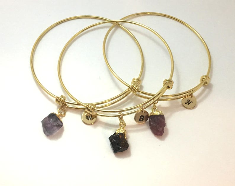 May include: Three gold bangle bracelets with charms. The charms are small, rough-cut gemstones in purple, red, and black. Each bracelet has a small gold disc with a letter engraved on it: N, W, and B.