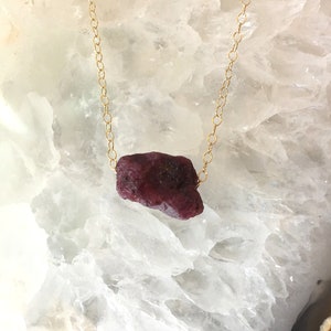 May include: A gold chain necklace with a raw ruby pendant. The ruby is a deep red color and has a rough, natural texture.
