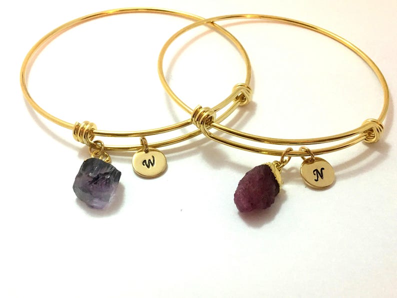 May include: Two gold bangle bracelets with charms. One charm is a purple crystal and the other is a red crystal. Each charm has a small gold disc with a letter on it. The letter on the purple crystal charm is "W" and the letter on the red crystal charm is "N".