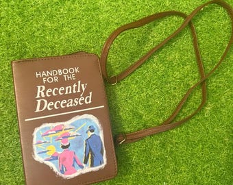 May include: A brown crossbody bag with the text "Handbook for the Recently Deceased" on the front. The bag features a colourful illustration of two figures. The strap is brown and adjustable. The bag is lying on a green surface.