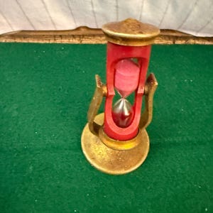 May include: A vintage-style hourglass featuring a red plastic frame and a gold-toned metal base and top. The hourglass is mounted on a swiveling gold-toned metal stand and contains pink sand that is currently flowing into the bottom glass bulb.