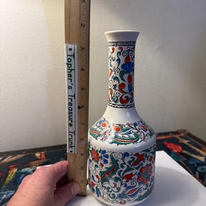 May include: A white ceramic vase with a narrow neck and a rounded base, decorated with a colorful floral pattern in red, blue, and green. A wooden ruler is placed next to the vase, showing a height of approximately 11 inches.