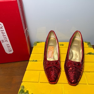May include: A pair of red sequined shoes with a small bow on the toe, featuring a gold interior lining. The shoes are displayed on a yellow brick-patterned base next to a red rectangular box with the text THE RUBY SLIPPERS printed on the side.