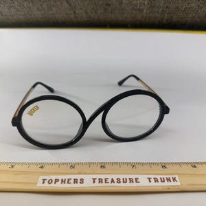 May include: Black-rimmed eyeglasses with a unique oval shape and gold-colored temples. The word "WICKED" is printed in gold on the left lens. The glasses are set against a white background.