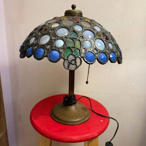 May include: A table lamp with a dome-shaped stained glass shade featuring iridescent white, blue, and green glass cabochons set in a dark metal frame. The lamp has a simple brass-toned metal base and a black power cord with a pull chain switch.