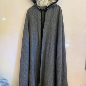 May include: A gray hooded cloak with a black trim and a toggle closure. The cloak is made of a textured fabric and hangs from a gold hanger. The hood is lined with a lighter-colored fabric.