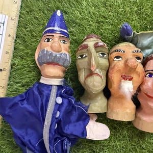 May include: Four hand puppets with carved wooden heads and painted facial features. One puppet wears a blue uniform with a matching hat. A wooden ruler measuring 10 inches is placed vertically next to the puppets for scale reference.