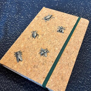 May include: A cork notebook with a green elastic closure. Five silver insect charms are attached to the cover: a bee, a cicada, two beetles, and a fly.